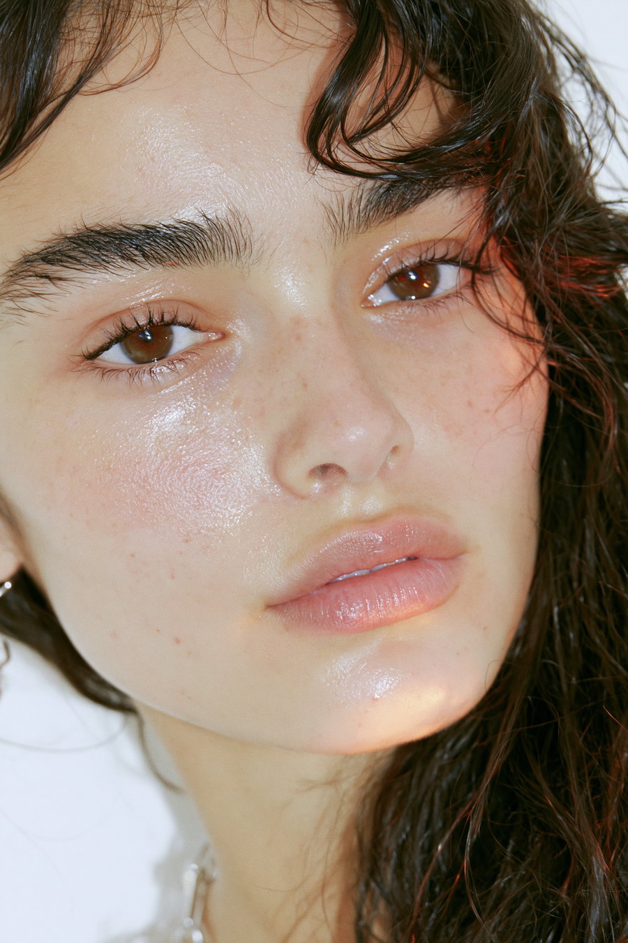 A close-up portrait of a young woman with natural skin tones, thick dark brows, almond-shaped brown eyes, and full lips, featuring glossy skin and a soft expression.
