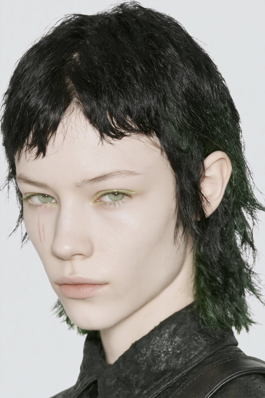 A close-up portrait of a pale young woman with messily styled short black and green hair, showcasing a natural skin texture, light gray background, and a subtle focus on her striking makeup.