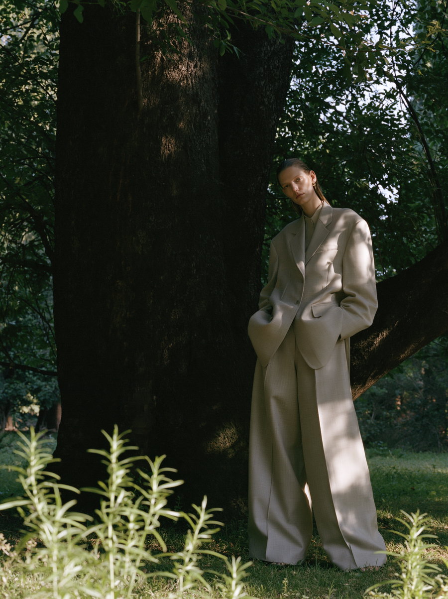 A full-length portrait of a woman in a structured oversized blazer and loose trousers, standing under a large tree in a lush woodland. Soft sunlight creates shadows and highlights on her outfit and the grass, with a blurred background providing depth.