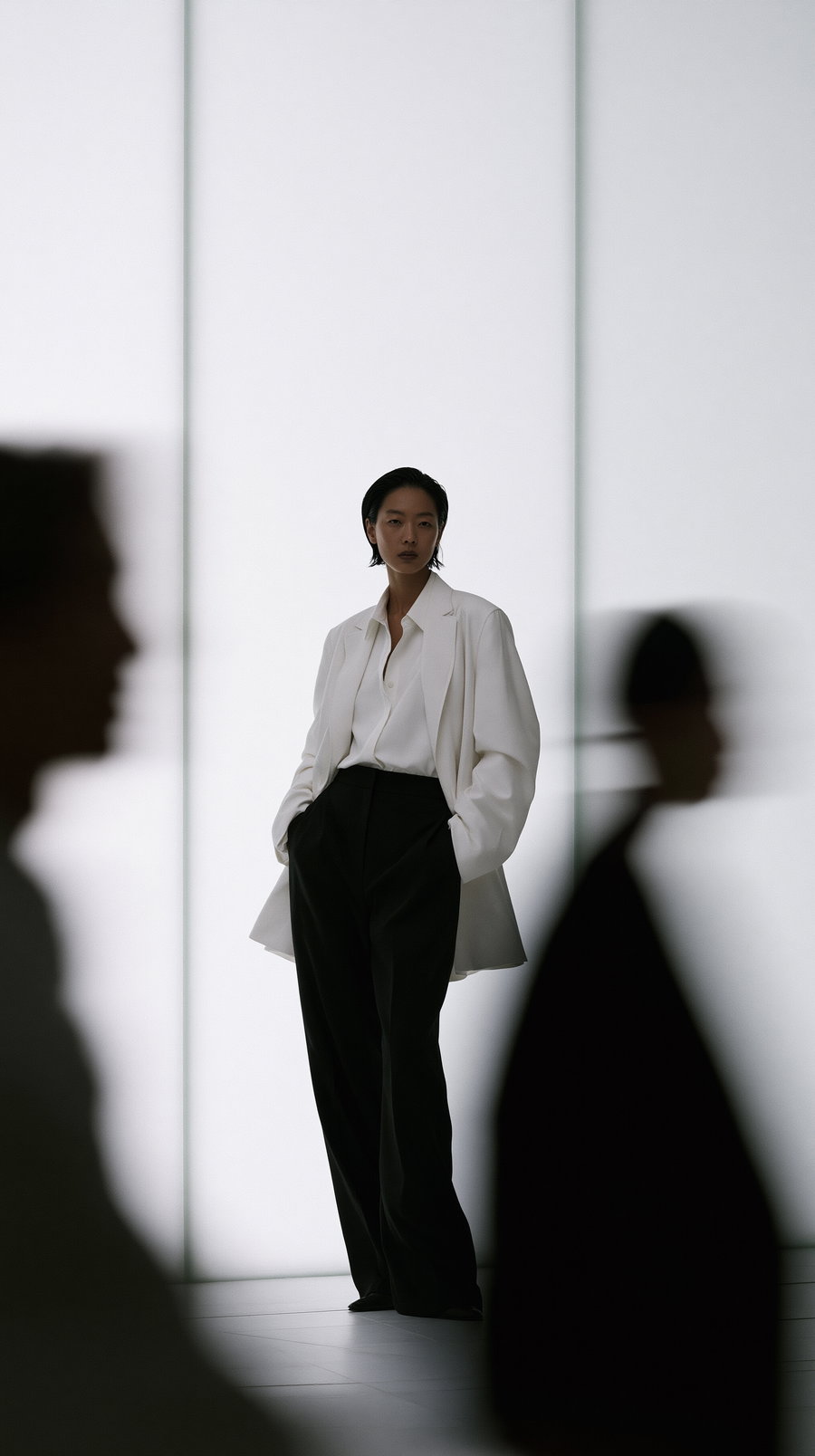 An elegant Japanese woman in a white outfit stands against a white vertically striped glass wall. Blurred figures fill the foreground, creating a dynamic, minimalist scene.