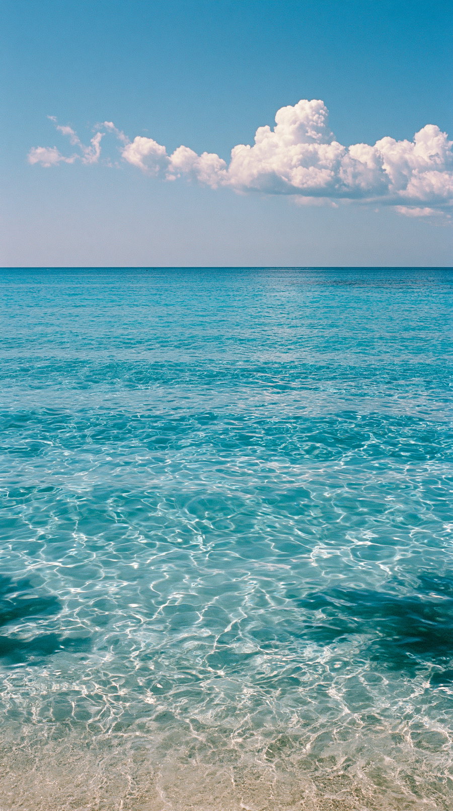 A serene view of a turquoise sea meeting a clear blue sky with soft white clouds and gentle waves reflecting the sunlight.