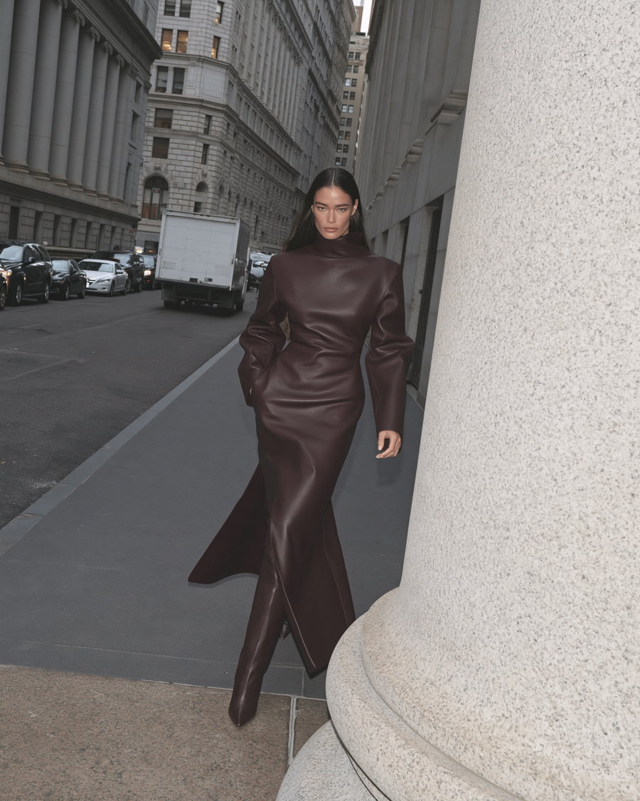 A confident model in a burgundy leather dress walking on a quiet city street lined with neoclassical buildings, featuring a large granite column in the foreground.