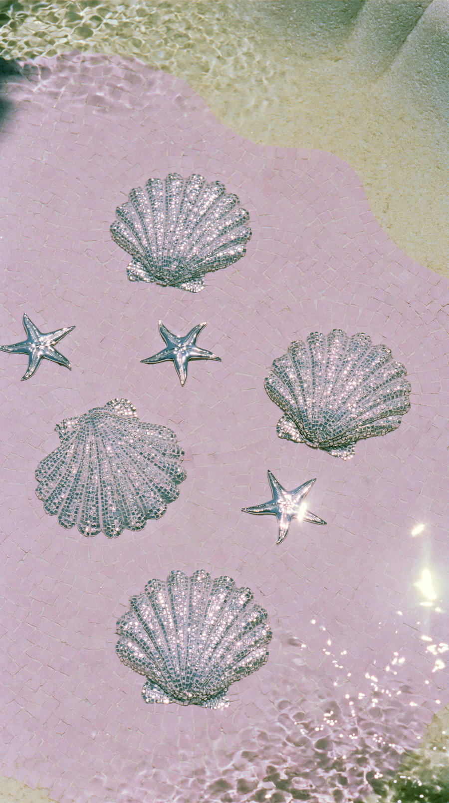 Flat lay view of a shallow pink stone fountain with silver seashell and starfish mosaics on the floor, reflected in clear water.