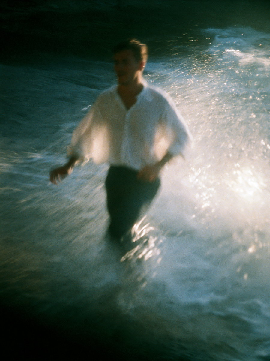 A man in a flowing linen shirt walking through sparkling ocean water with blurred motion effects and a dreamy, grainy film texture.