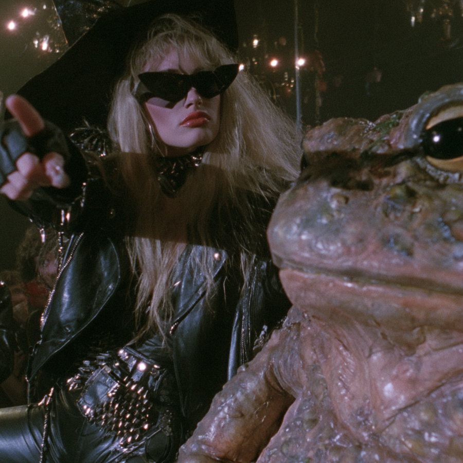 A woman in black leather and sunglasses points at the camera, standing next to a giant frog wearing sunglasses, in a dark, moody setting.