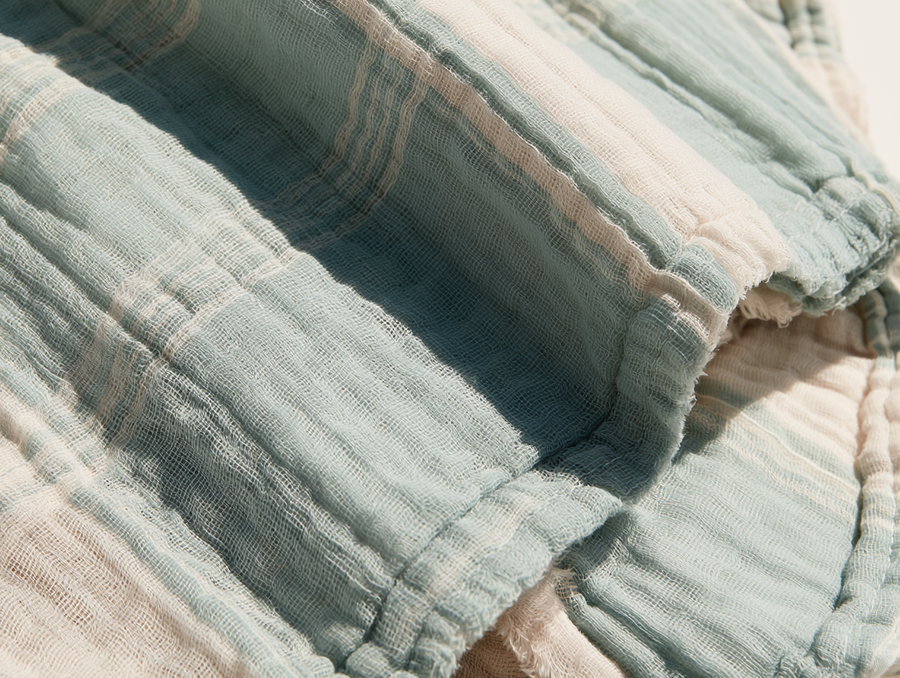 Dog bandana on a warm white surface, faded dusty blue and warm ecru colors, fine cotton gauze open-weave mesh, minimal fraying, sharp shadows from harsh sunlight.