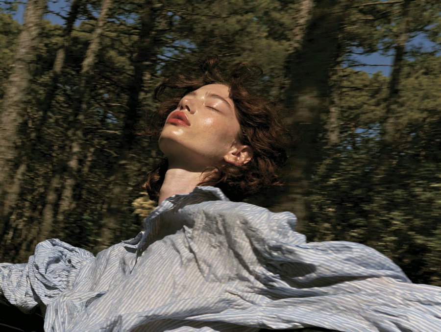 A young woman with brown hair leans back with her eyes closed, wearing a light blue and white striped shirt. The background features blurred green tree trunks and leaves, suggesting motion from a vehicle, creating a serene and dreamlike atmosphere.