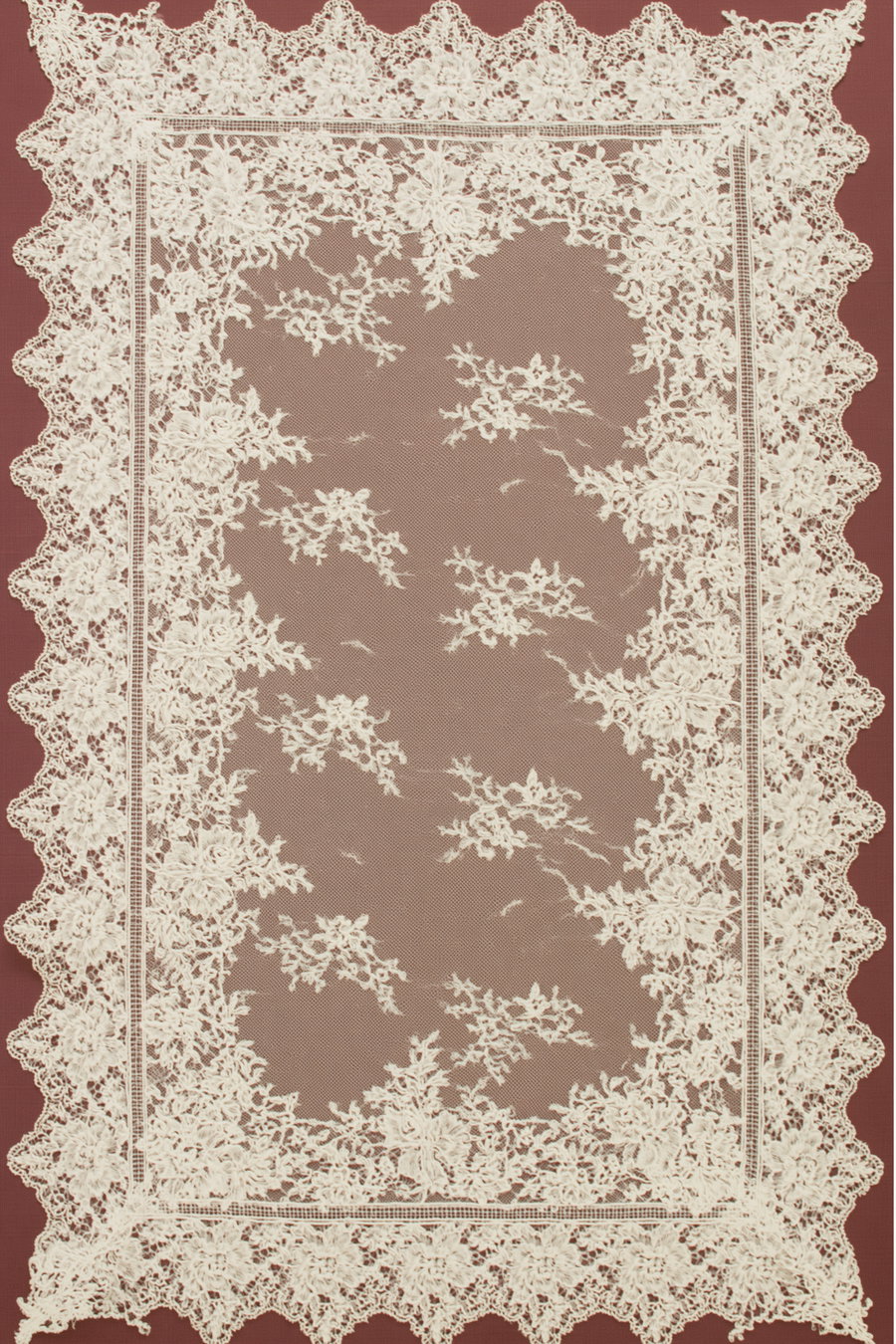 A square piece of white floral lace fabric with scalloped edges laid on a black studio surface. The lace features embroidered flowers and a sheer net center that allows the black background to show through, creating a high-contrast effect.