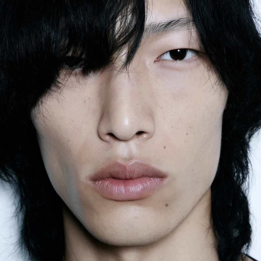 A close-up image of a young Japanese man with slightly wavy black hair and bangs, featuring flawless porcelain skin, deep brown eyes, and rosy lips, set against a white studio background.