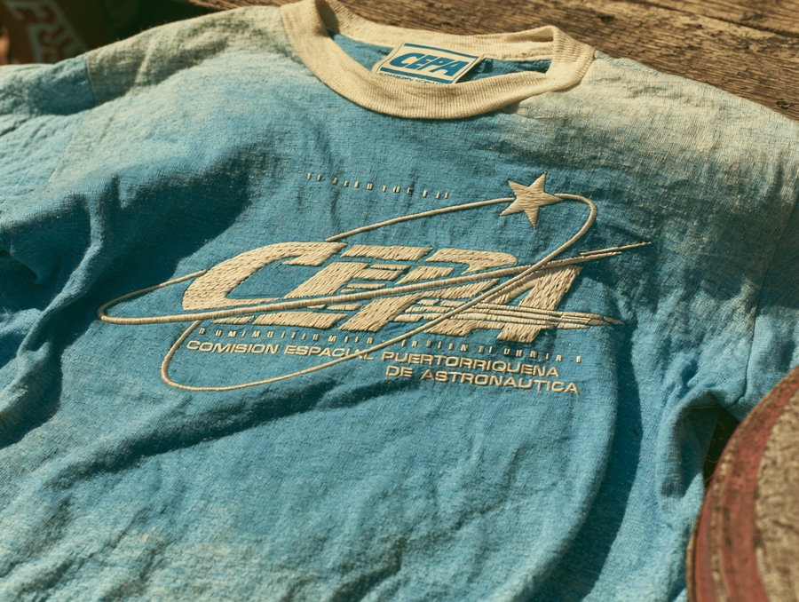 A vintage T-shirt featuring the embroidered CEPA logo in cream on cobalt blue, with an elliptical orbit line and a five-pointed star. The text 'COMISION ESPACIAL PUERTORRIQUENA DE ASTRONAUTICA' is displayed beneath in small monospaced font.
