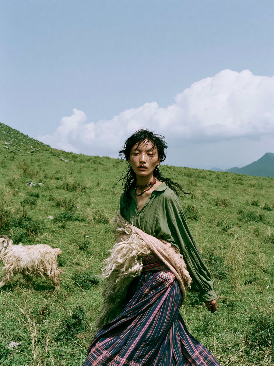 A young woman with flowing dark hair stands in a grassy field, wearing a green top and a colorful, striped skirt. She is surrounded by a bright blue sky with fluffy clouds and a hilly landscape. A sheep is visible nearby, enhancing the serene countryside atmosphere.